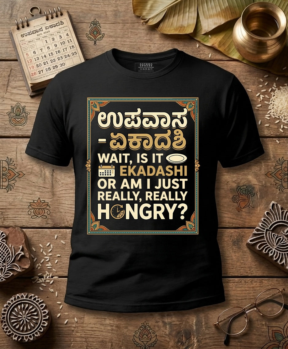 A black short-sleeve graphic T-shirt laid flat on a rustic wooden surface with decorative props including a calendar, brass pot, glasses, and carved ornaments. The shirt features a large rectangular printed design with Devanagari or Kannada text at the top and English text below reading, 'WAIT, IS IT EKADASHI OR AM I JUST REALLY, REALLY HUNGRY?'.